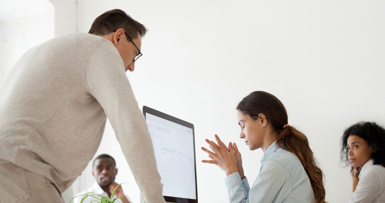 Boss-pointing-finger-while-speaking-to-seated-intern-at-desk