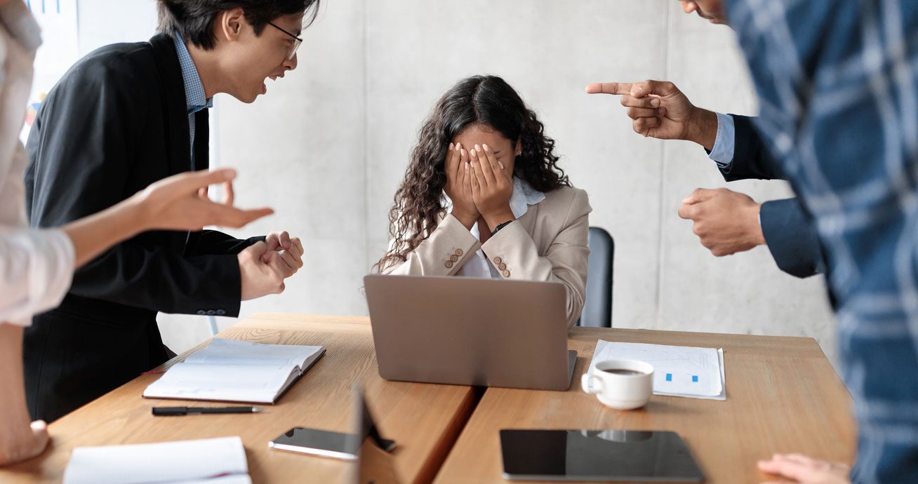 Coworkers-yelling-at-seated-woman-covering-face-at-desk