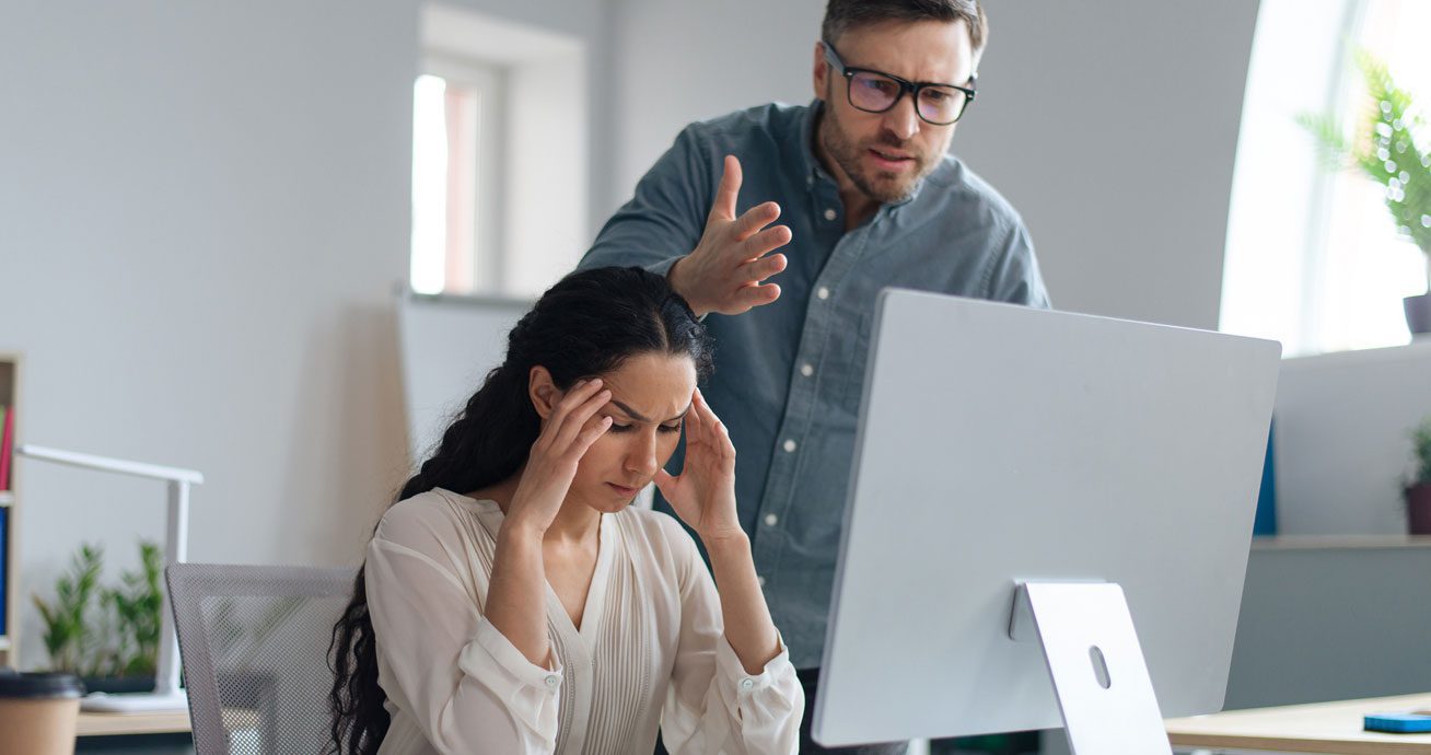 Male-boss-pointing-and-speaking-to-seated-female-employee-at-desk