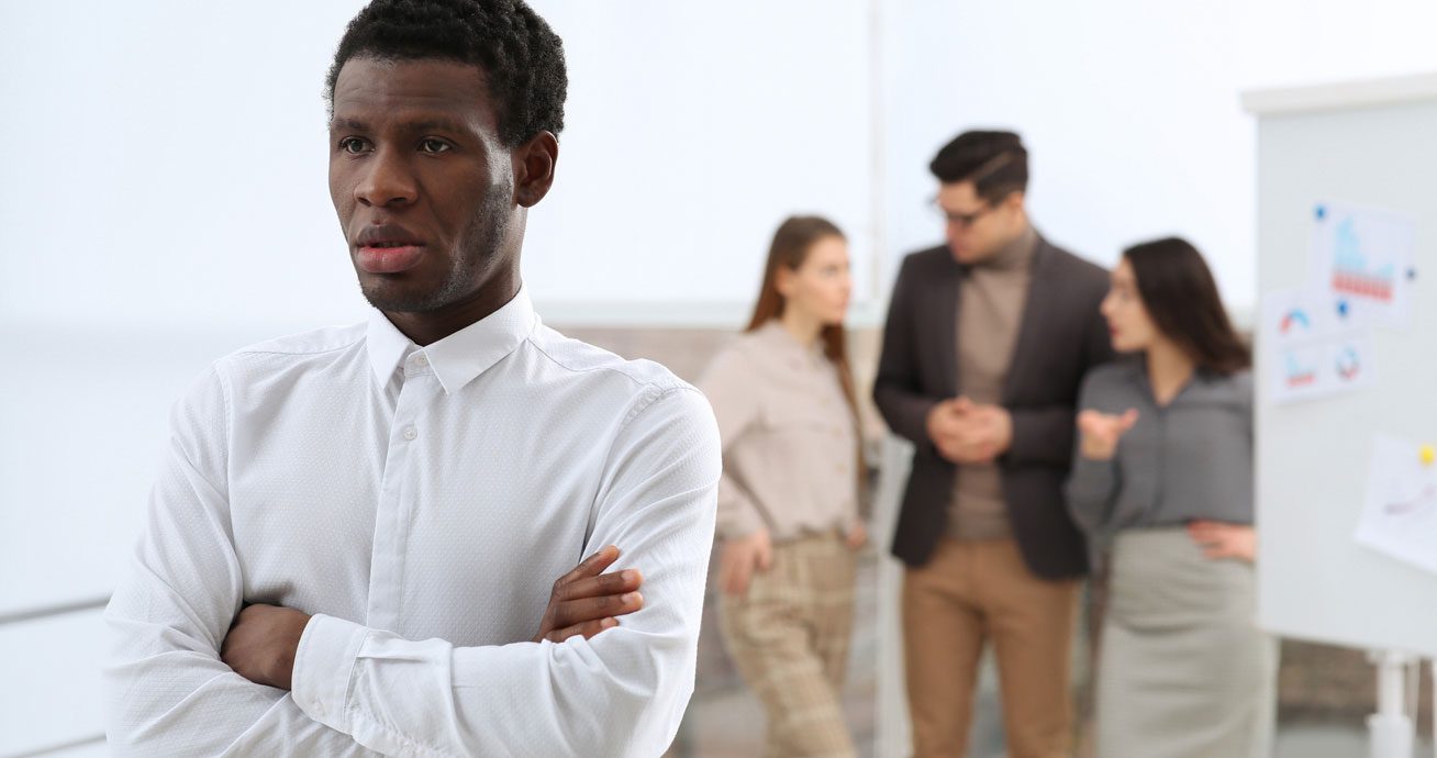 Man-sitting-alone-at-desk-while-coworkers-talk-nearby