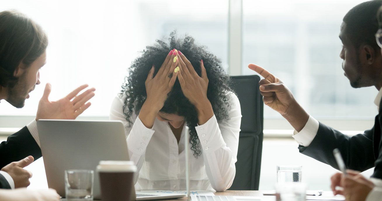 Woman-sitting-at-desk-while-coworkers-point-fingers-toward-her