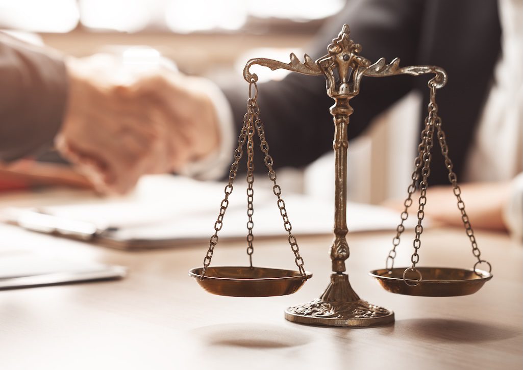 brass-scales-of-justice-on-a-desk-with-two-people-shaking-hands-behind