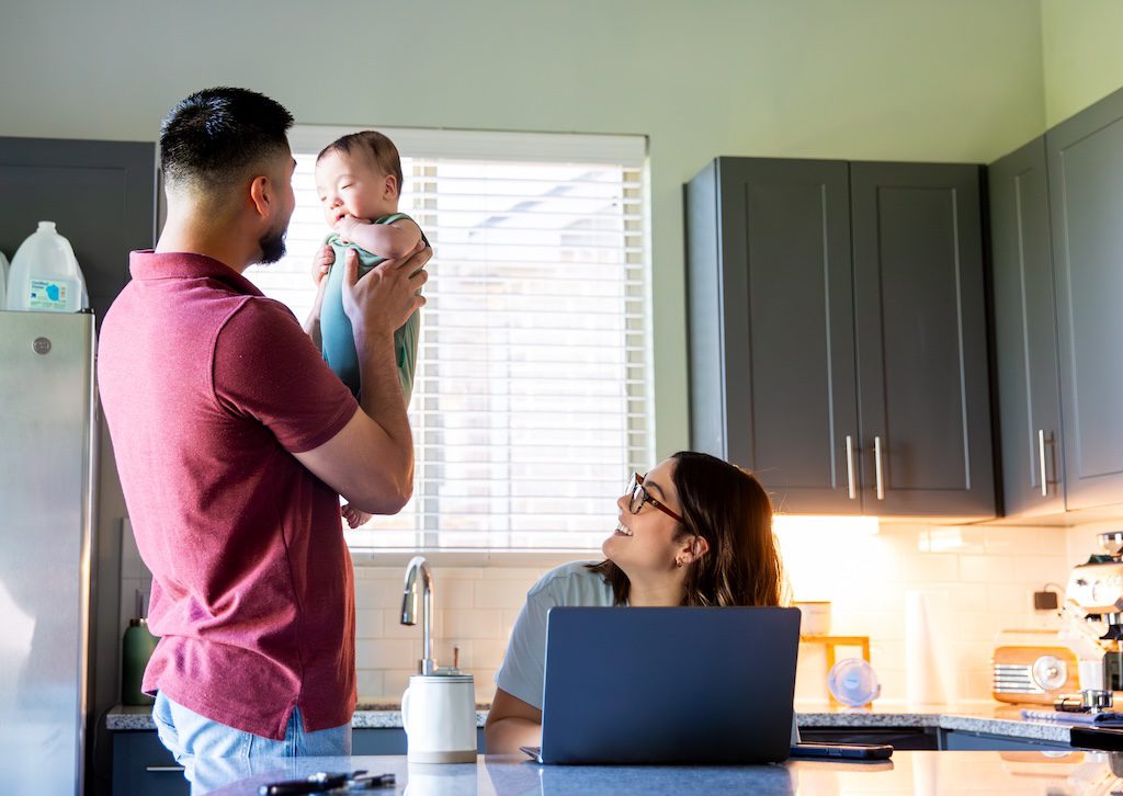 man-holding-a-baby-in-a-kitchen-while-a-woman-works-on-a-laptop