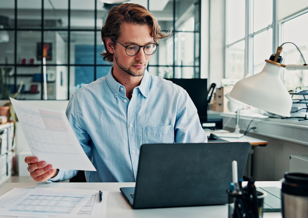 man-reviewing-a-document-at-a-desk-with-a-laptop-and-paperwork