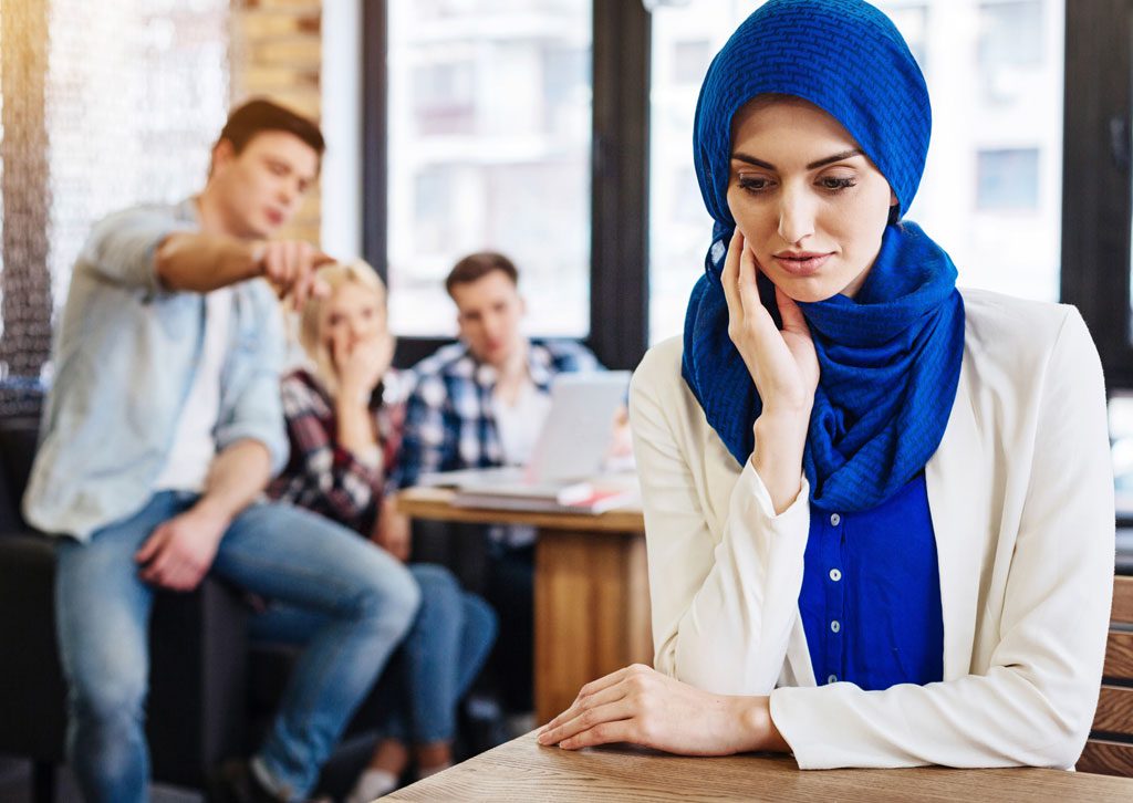 woman-in-a-headscarf-sitting-apart-from-a-group-of-coworkers-talking-behind-her