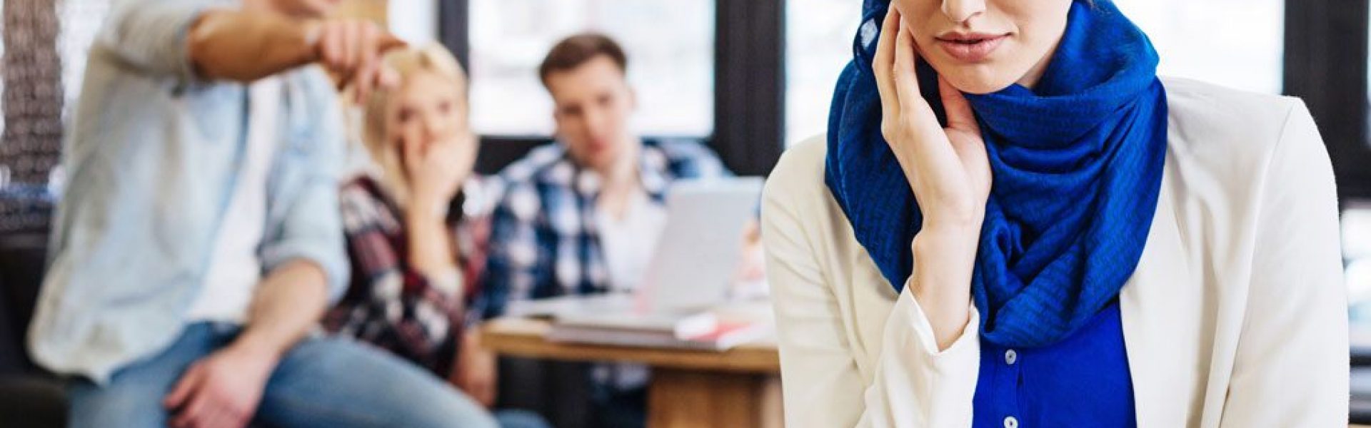woman-in-a-headscarf-sitting-apart-from-a-group-of-coworkers-talking-behind-her